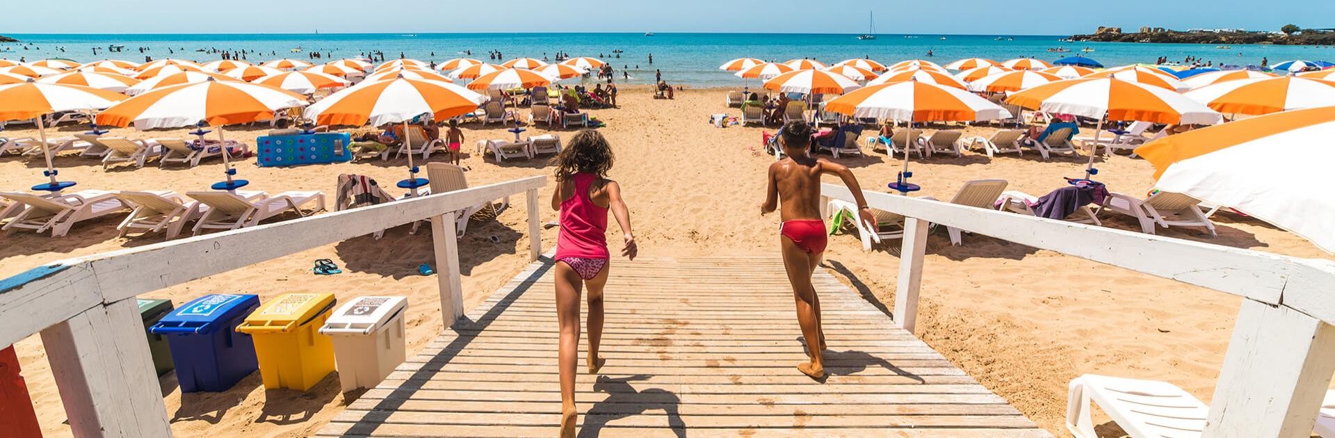 Plage bondée avec parasols orange et blancs, enfants courant vers la mer.