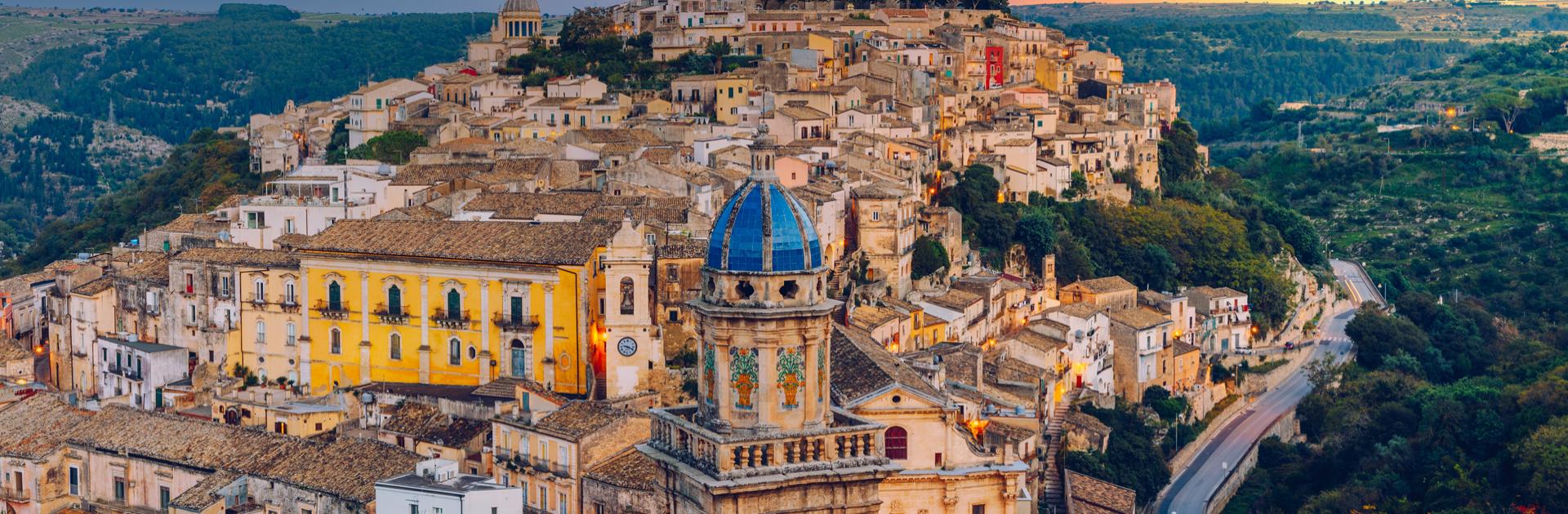 Blick auf Ragusa mit historischen Gebäuden und hügeliger Landschaft bei Sonnenuntergang.